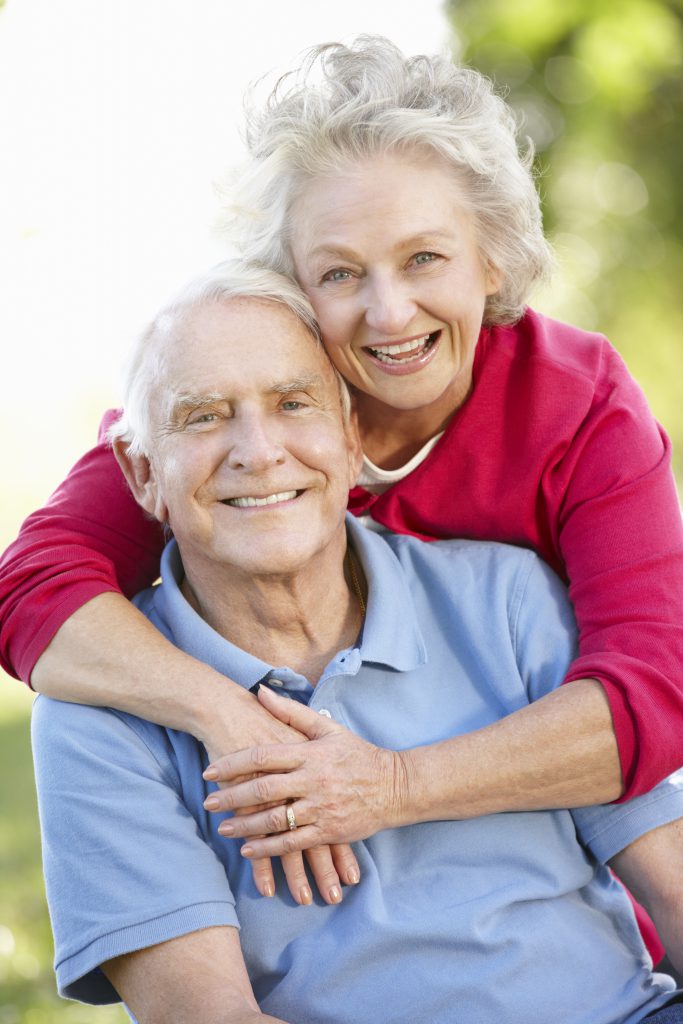 older couple smiling outside together as she puts her arms around him